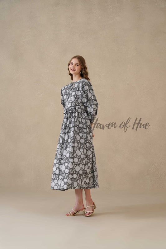 A woman standing in a charcoal black and cream floral cotton midi dress with full sleeves and round neck.